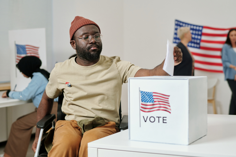 African American young man with disability sitting in wheelchair and putting ballot paper into box with USA flag standing on desk